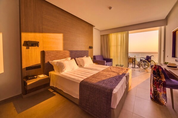 A hotel room with a large bed, a desk, a chair, a wheelchair, and a view of the sea.