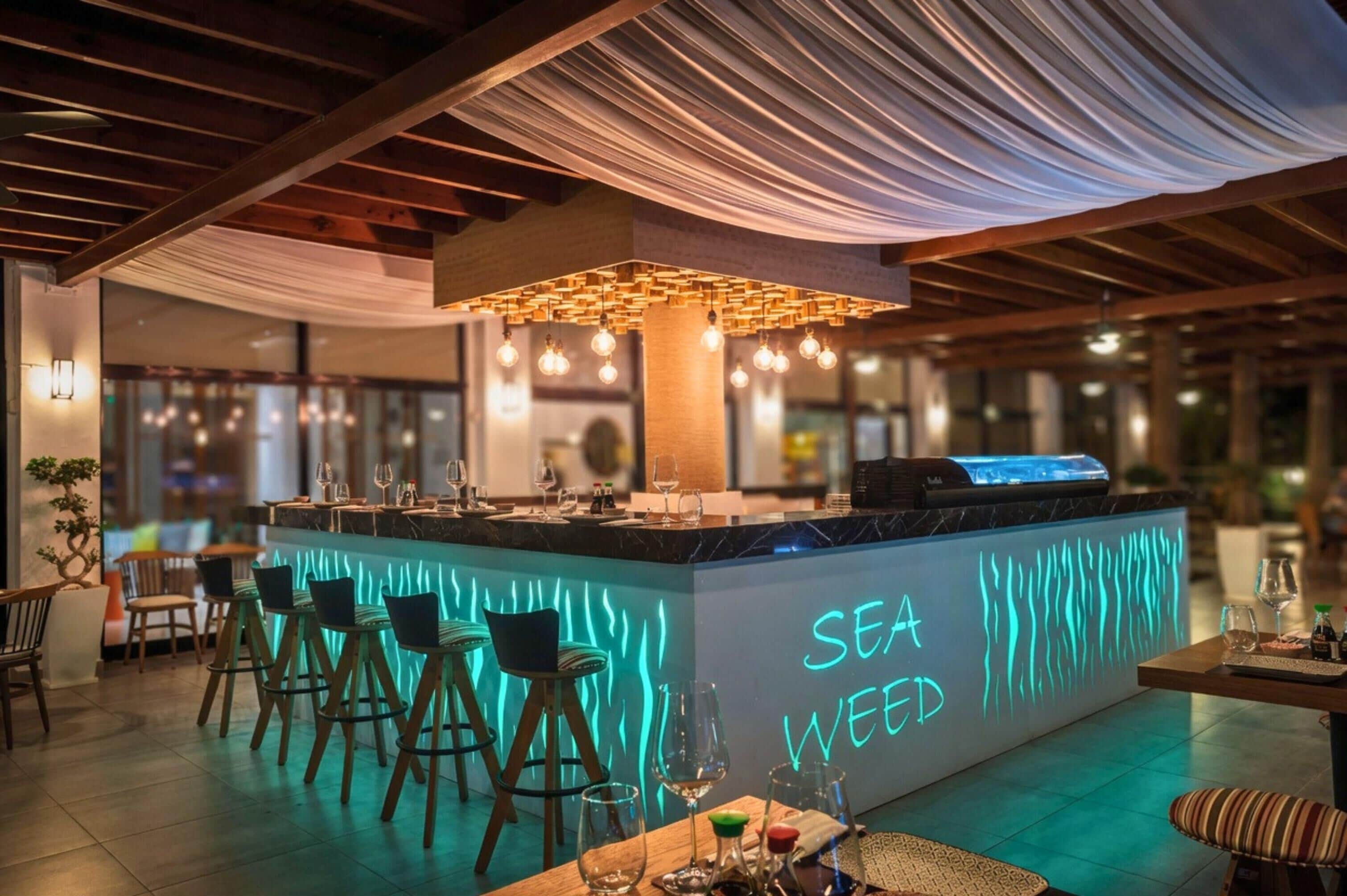 Una zona de bar con taburetes, un letrero de neón que dice 'SEA WEED' y una lámpara moderna.