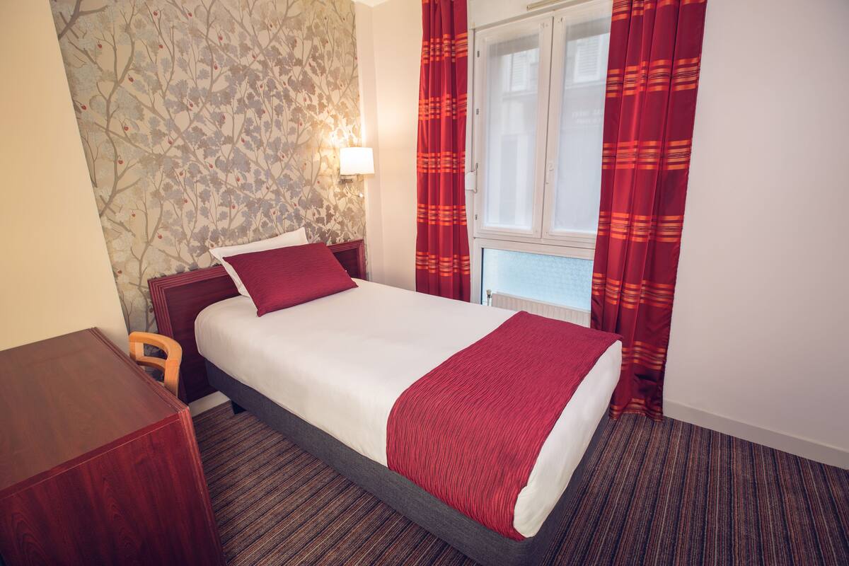 A hotel room with a bed, a wooden desk, a window with curtains, and a wall with a floral pattern.