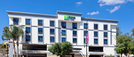 Un hotel Holiday Inn Express & Suites con un diseño moderno, varios estacionamientos y un cielo azul despejado.