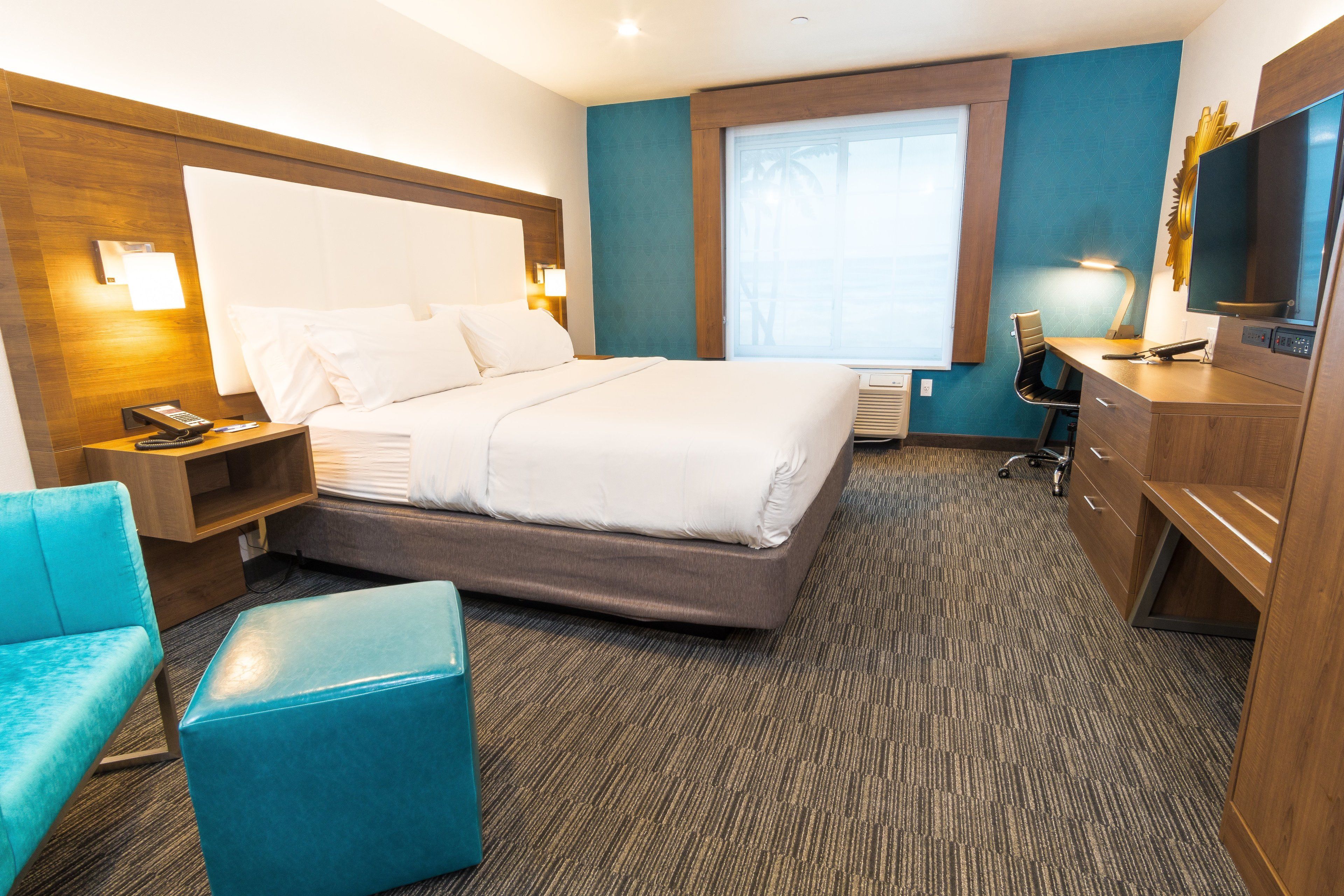A hotel room with a large bed, a desk with a chair, a TV, and a window with a view of palm trees.