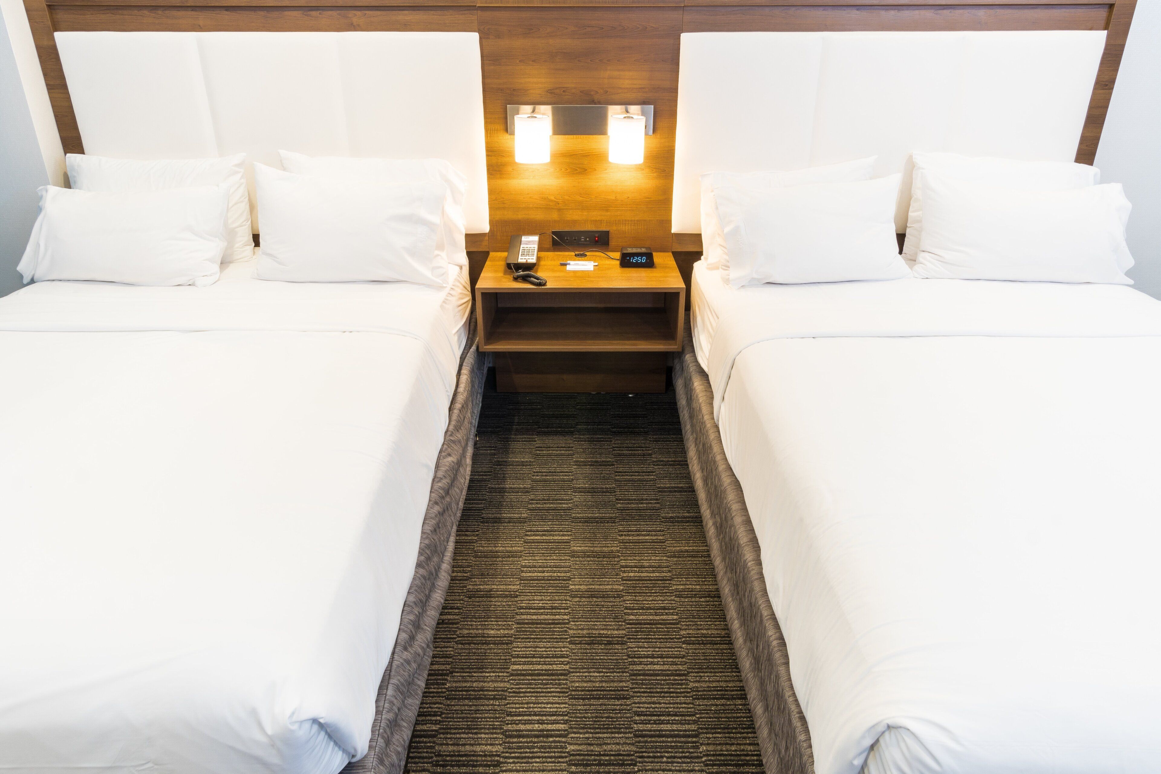 A hotel room with two beds, a nightstand with a phone and clock, and a carpeted floor.