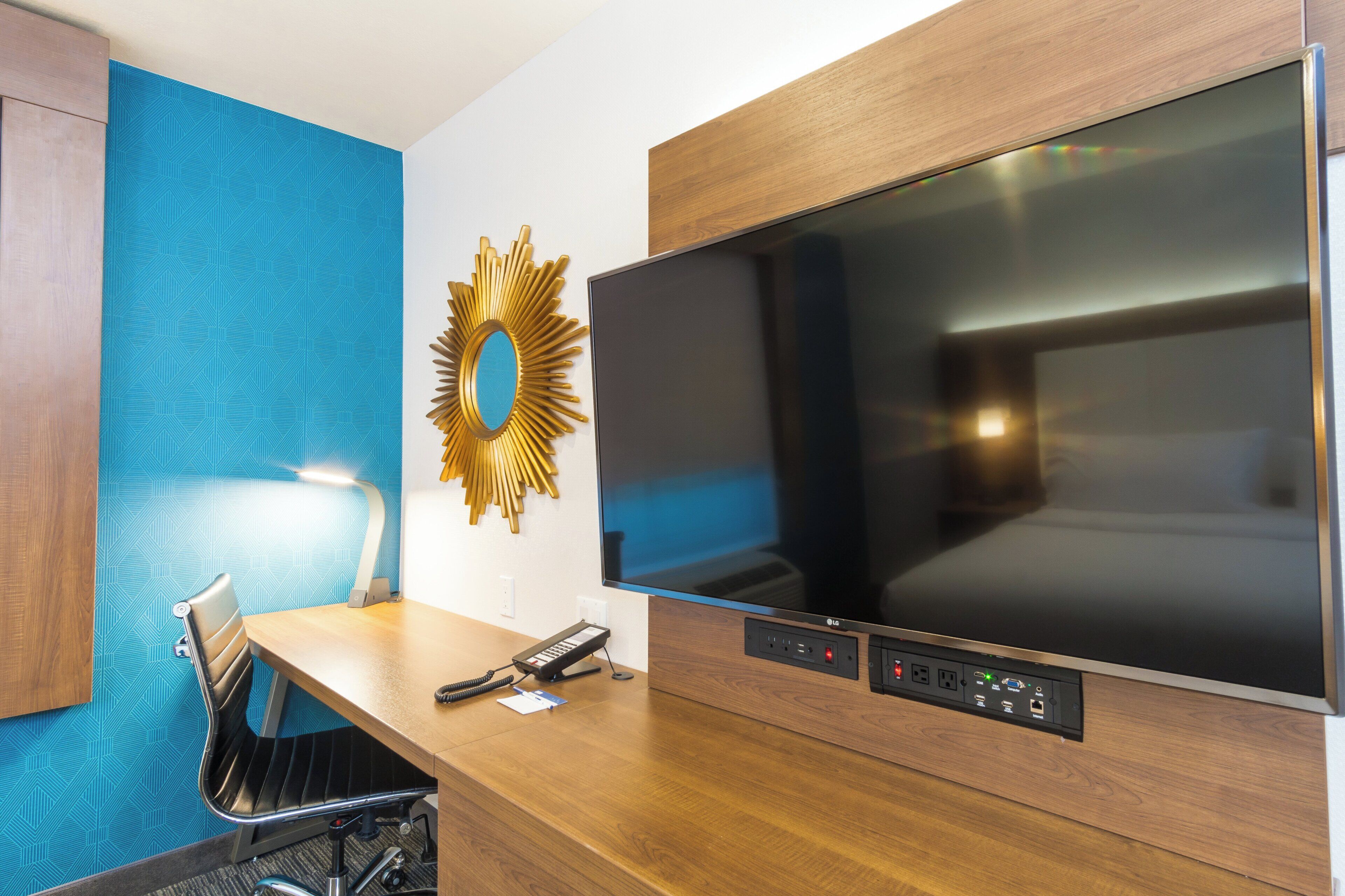 A hotel room with a large flat-screen TV, a desk with a telephone, and a chair.