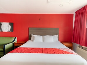 A hotel room with a bed, a green armchair, a nightstand, a red curtain, and a wall with a portrait. -  Rose Park Inn & Suites San Antonio (San Antonio)