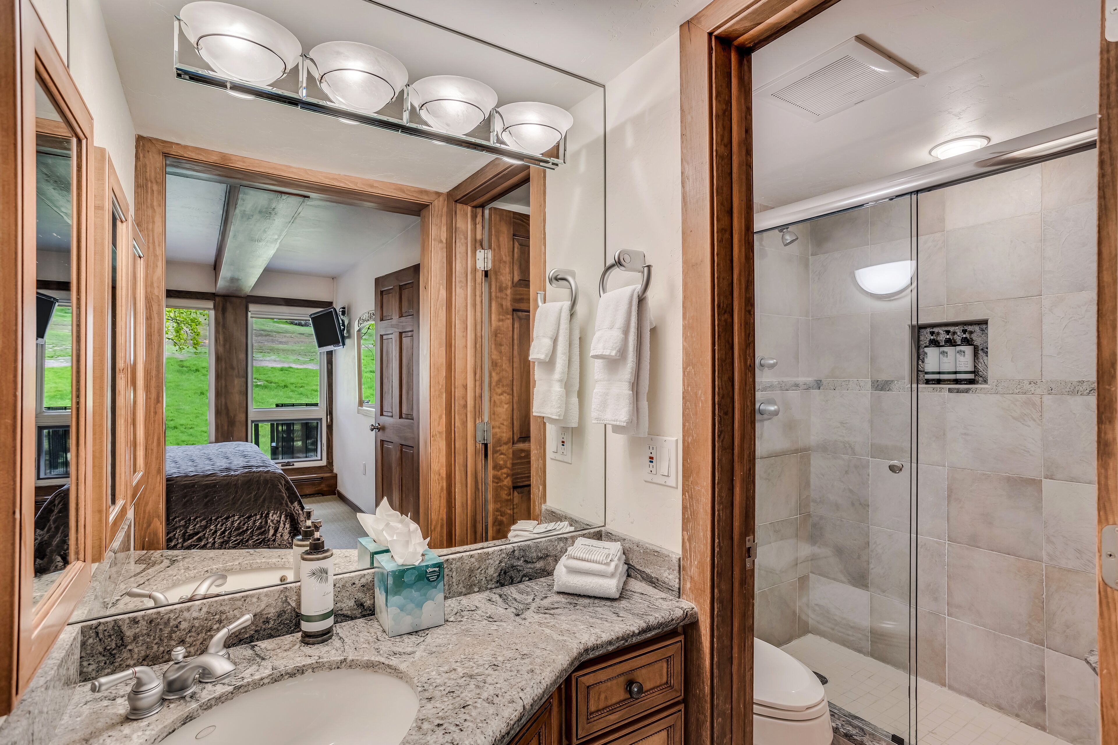 Un baño con un espejo grande, un lavabo con encimera de mármol, ducha con puertas de vidrio y vistas a una puerta de madera que da a un cuarto con cama.
