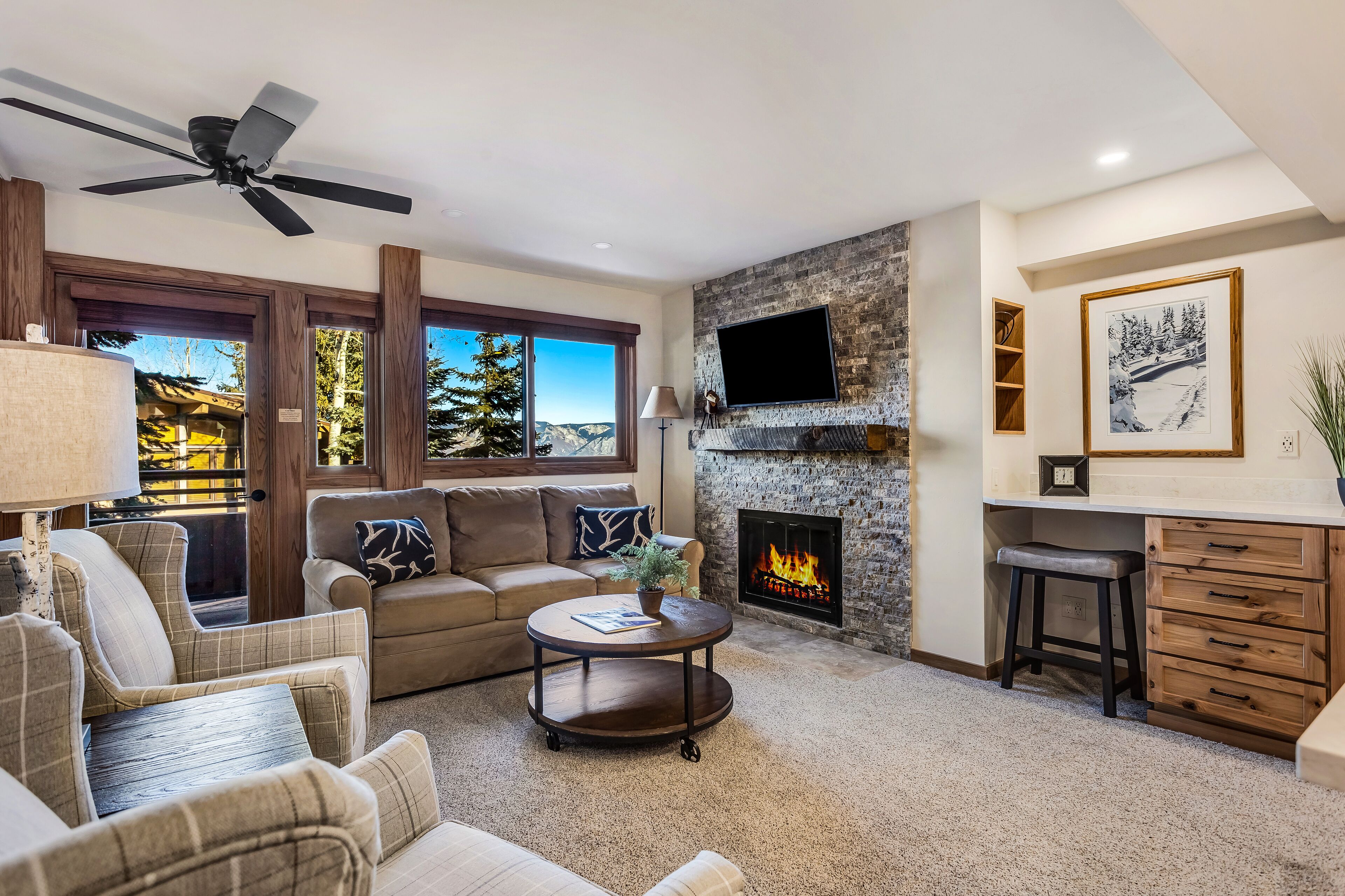 A cozy living room with a fireplace, a television mounted on a stone wall, and a ceiling fan.