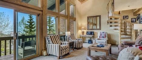 A spacious living room with large windows, a fireplace, and a wooden staircase.