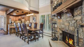 A spacious dining area with a stone fireplace, wooden beams, and a view of the kitchen.