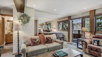 A spacious living room with a large couch, a coffee table, and a fireplace. There is a bedroom visible in the background with a large window.