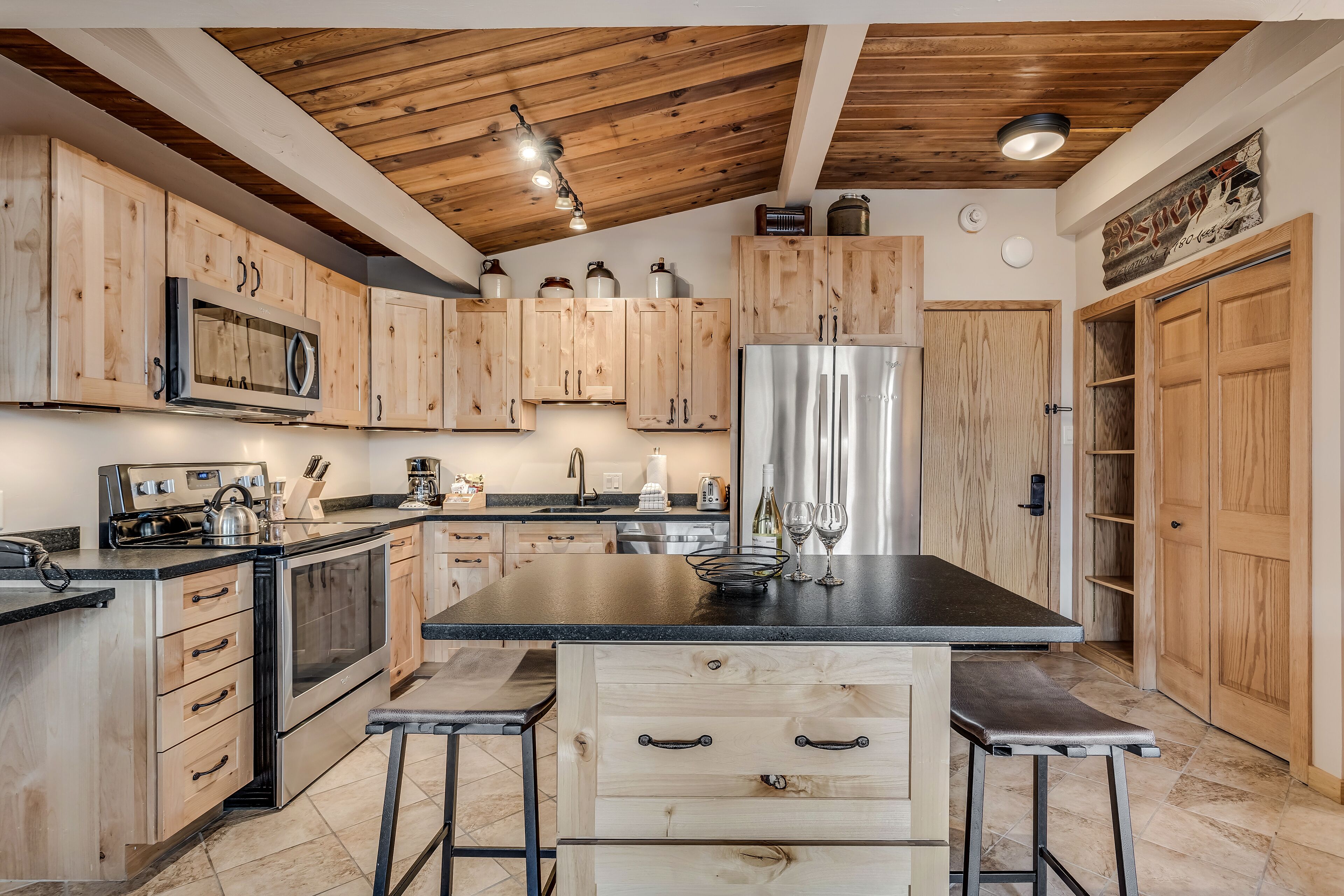 Una cocina con armarios de madera, una isla central con taburetes, electrodomésticos de acero inoxidable y techo de madera.