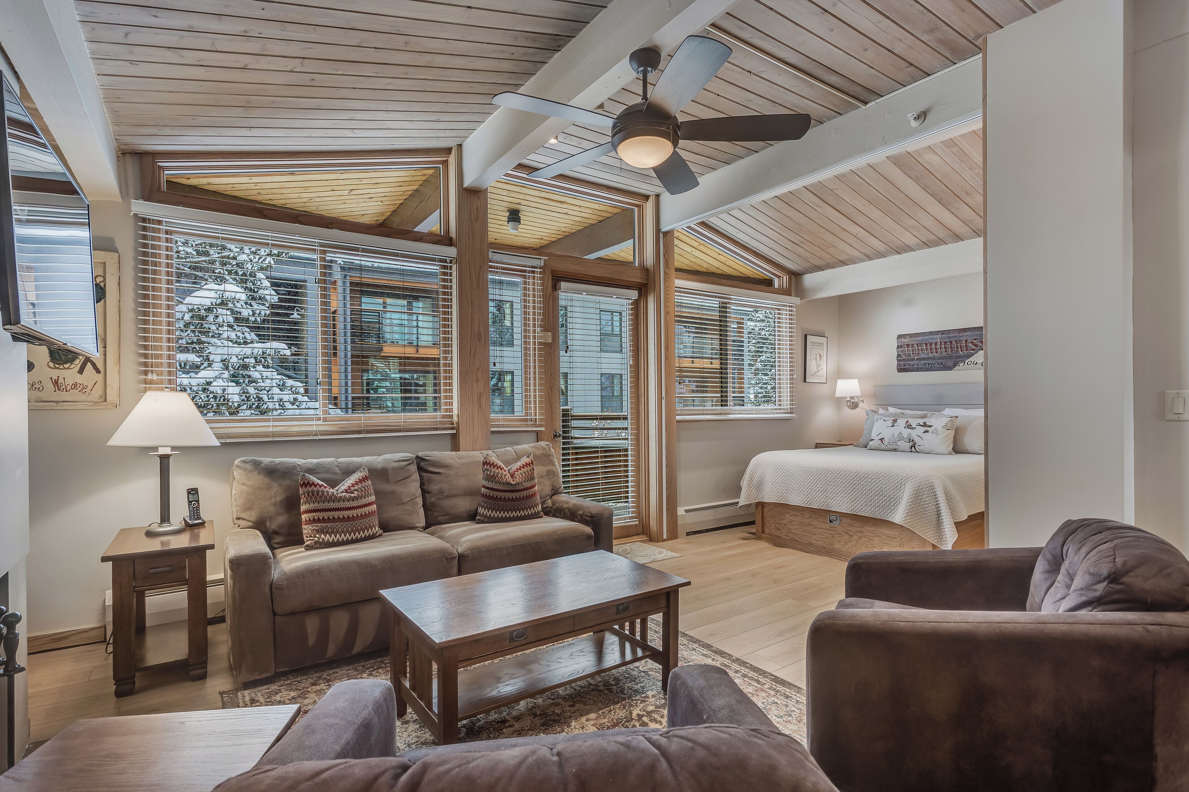 A spacious living room with a wooden ceiling, large windows with blinds, a sofa, a coffee table, and a bed with a white bedspread.