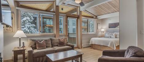 A spacious living room with a wooden ceiling, large windows with blinds, a sofa, a coffee table, and a bed with a white bedspread.