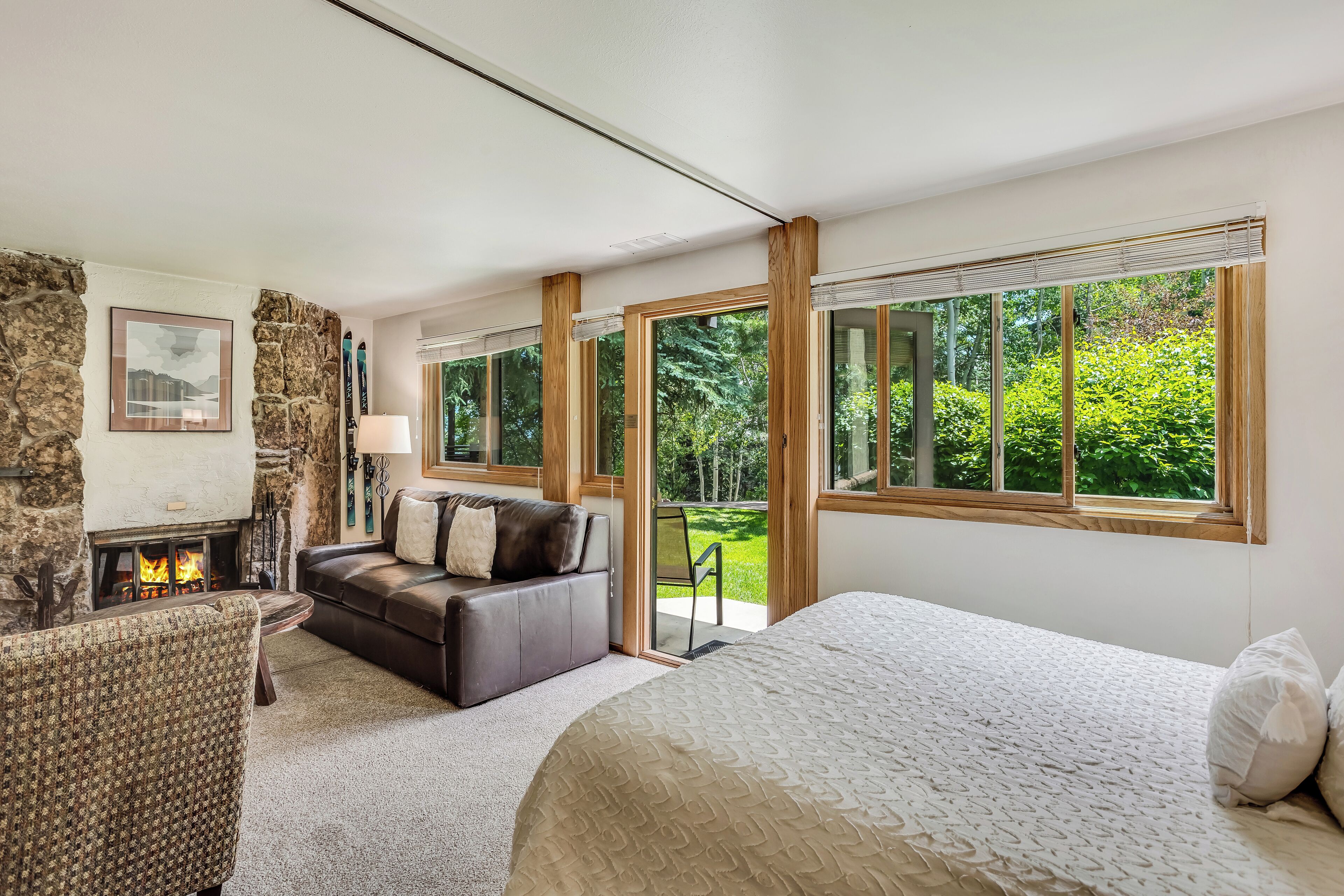 Une chambre spacieuse avec un grand lit, une cheminée, un canapé et une vue sur la verdure grâce à de grandes fenêtres.