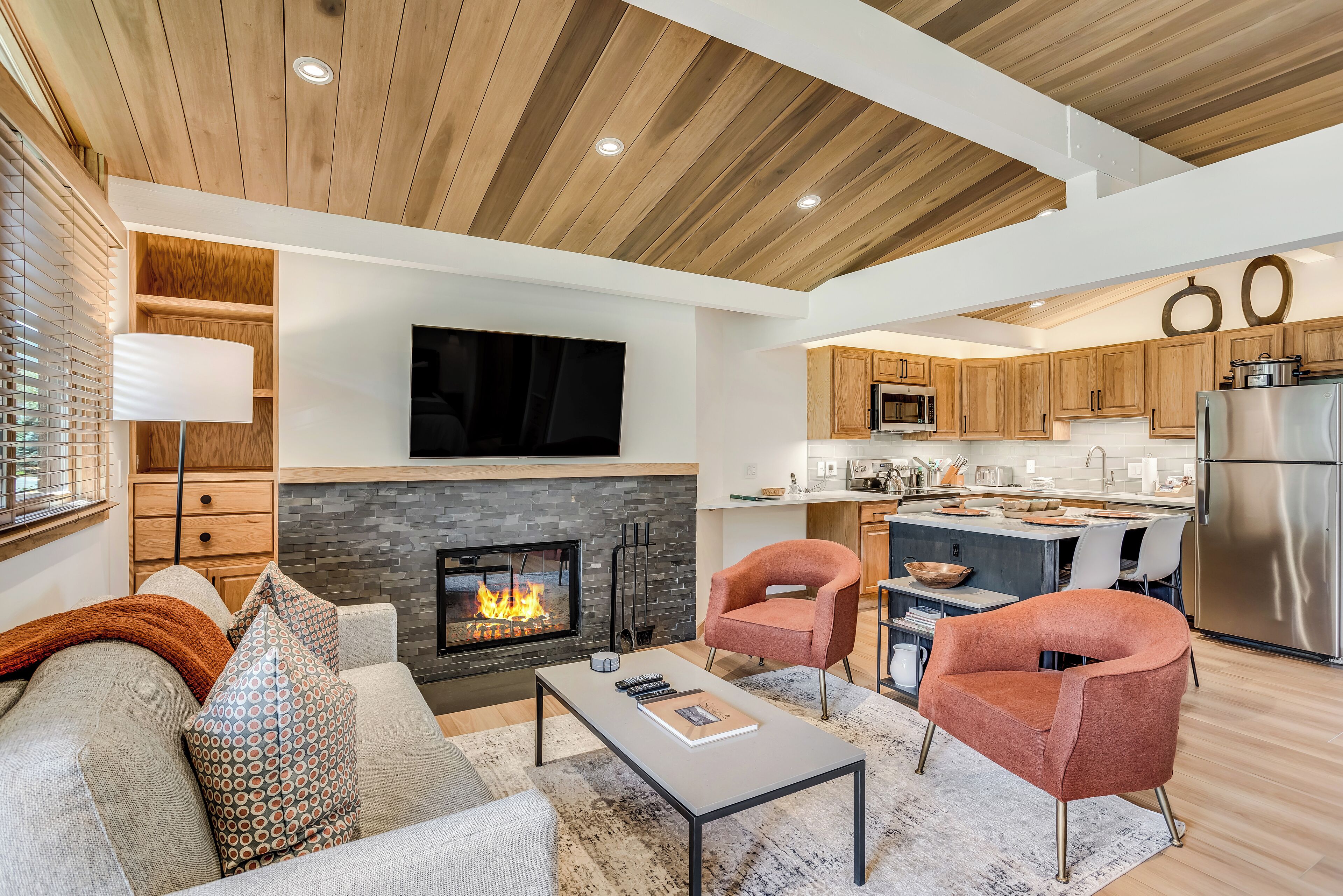 A modern living room with a fireplace, wooden ceiling, and a kitchen area with wooden cabinets and stainless steel appliances.
