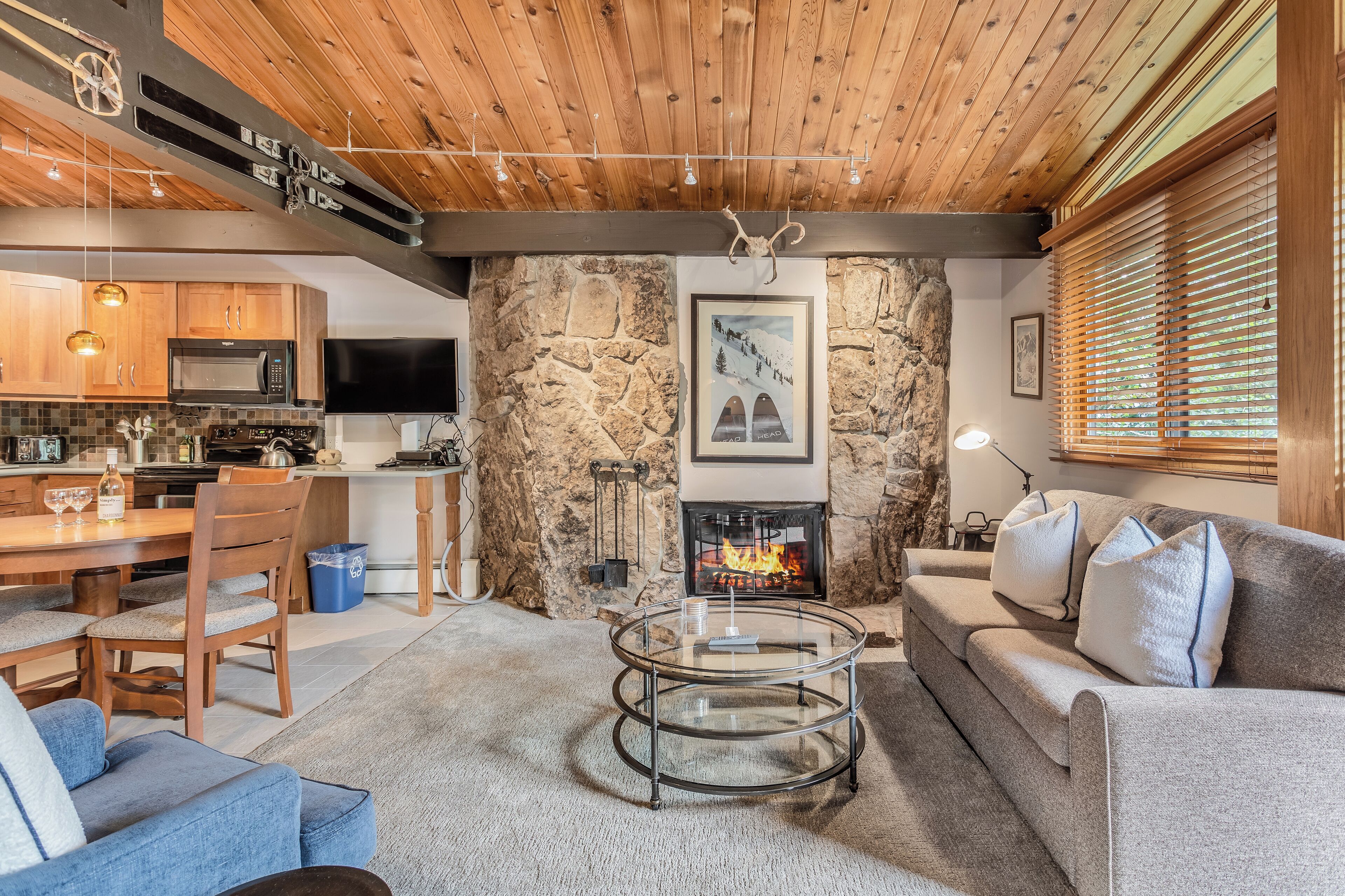 A cozy living room with a stone wall, a fireplace, a dining area with wooden furniture, and a kitchen with a modern design.