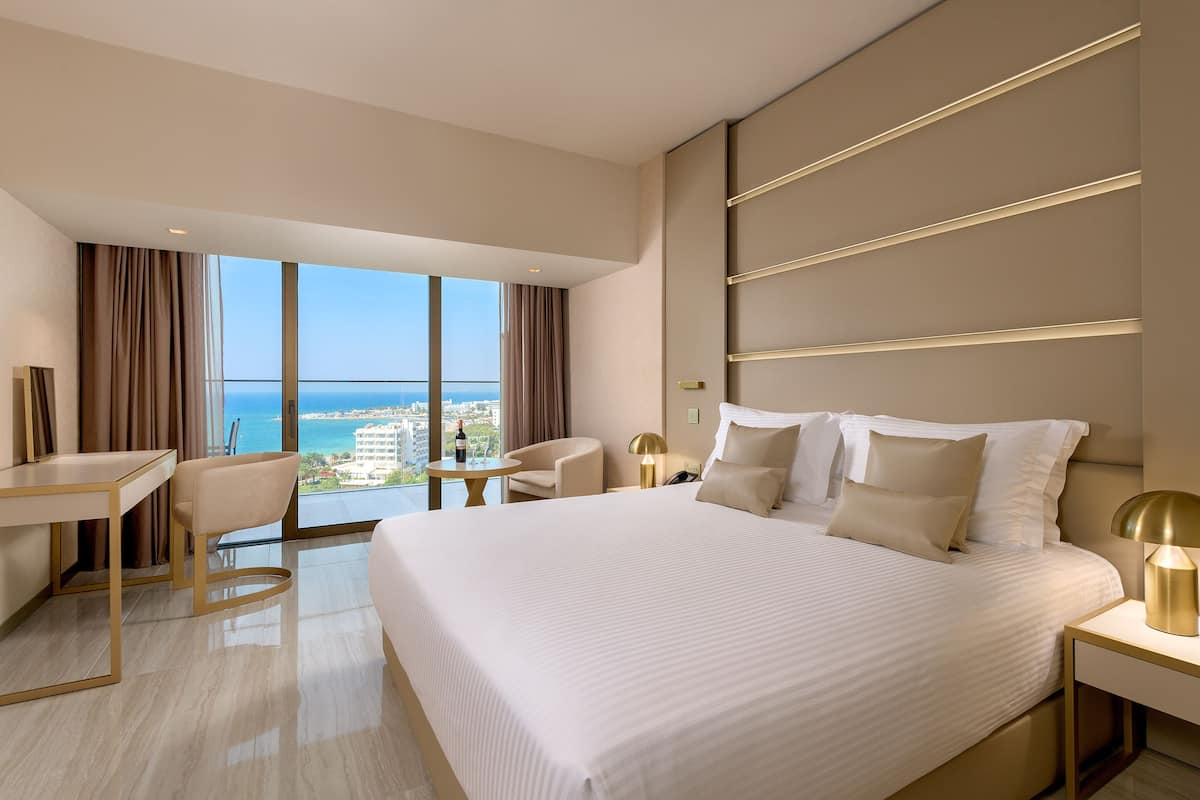 A modern hotel room with a large bed, a desk, a chair, and a view of the ocean.