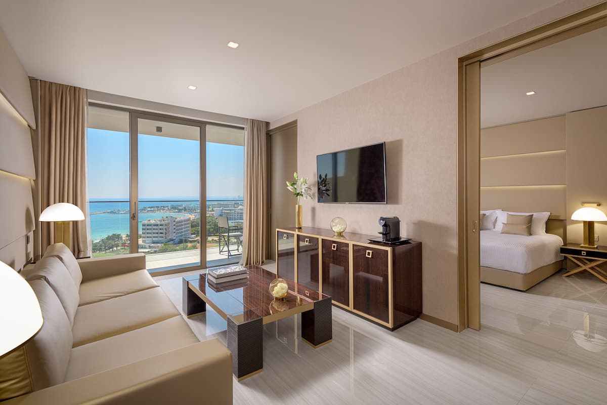 A modern hotel room with a large bed, a sofa, a TV, a desk, and a view of the ocean.