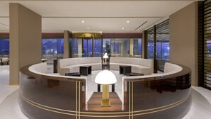A modern lobby with a curved reception desk, seating area, and large windows offering city views. - AMARANDE Adults Only (Ayia Napa)