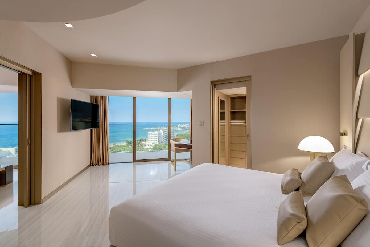 A hotel room with a large bed, a TV mounted on the wall, a desk, and a view of the ocean.