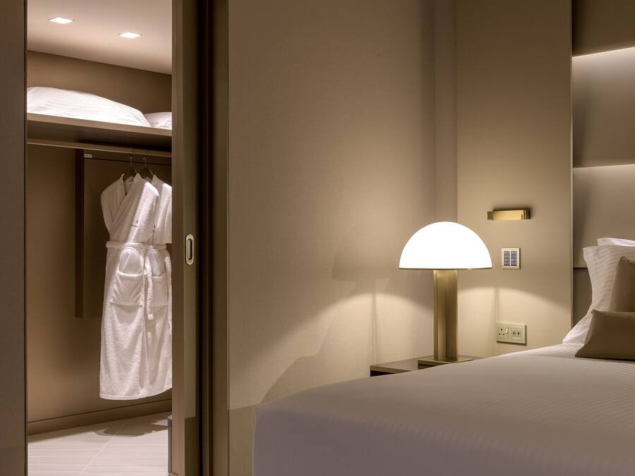 A hotel room with a bed, a bedside lamp, a robe hanging on a door, and a wall-mounted light fixture.