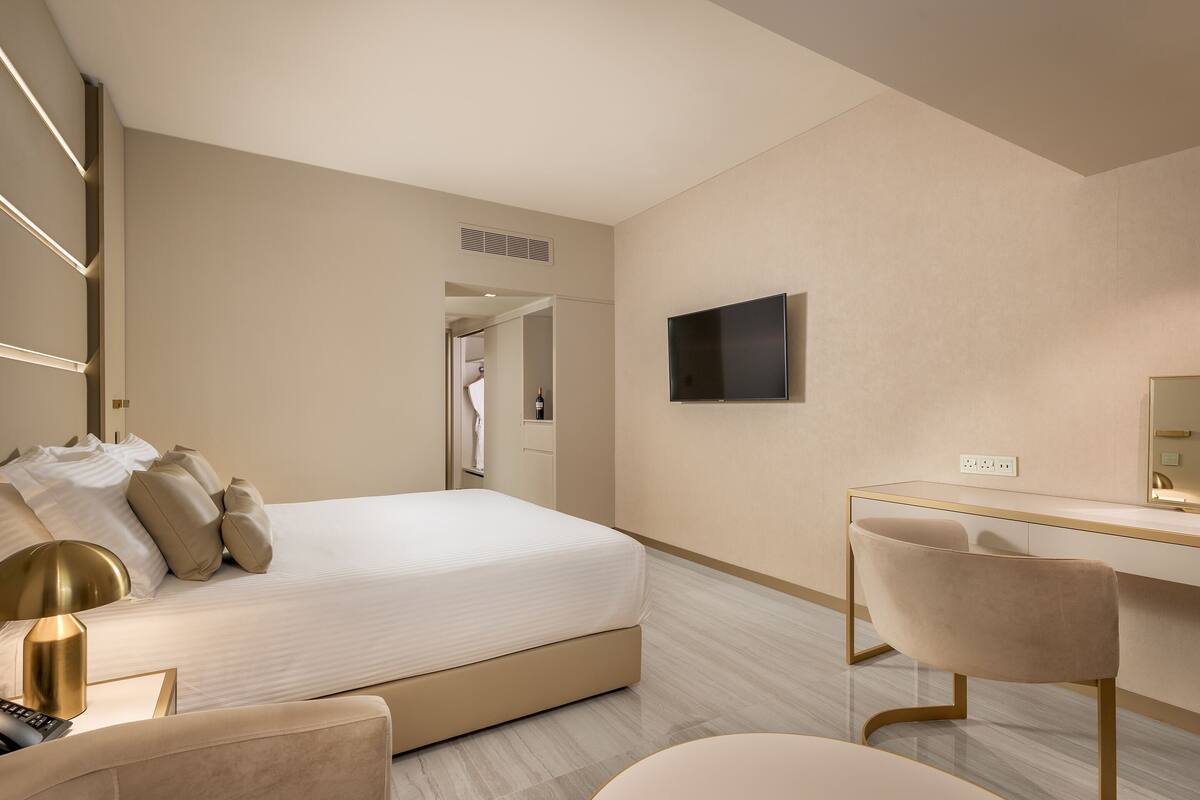 A modern hotel room with a large bed, a desk, and a chair. There is a television on the wall and a power outlet on the desk.