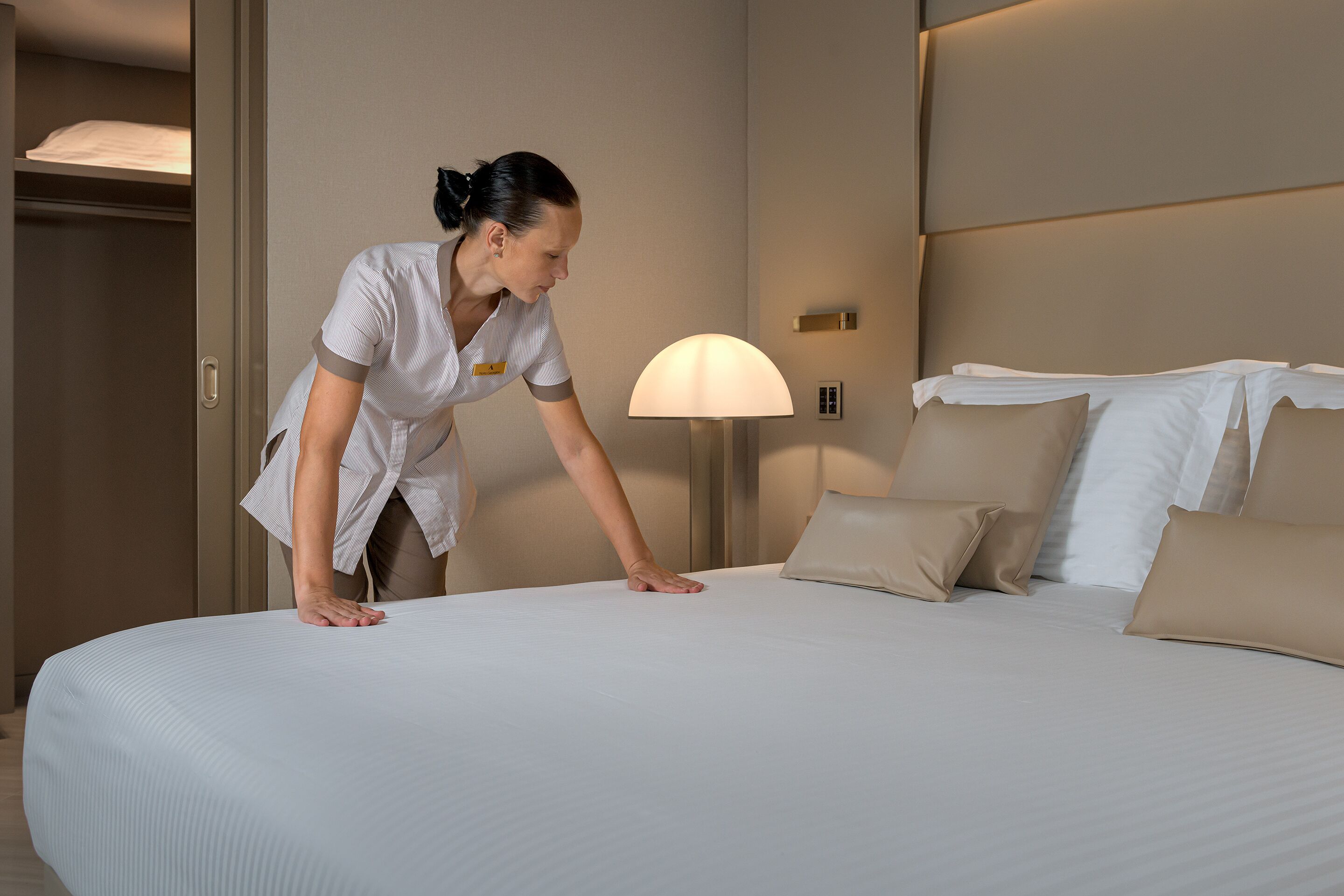 A neatly made bed with a white and beige color scheme, a bedside lamp, and a closed wardrobe.