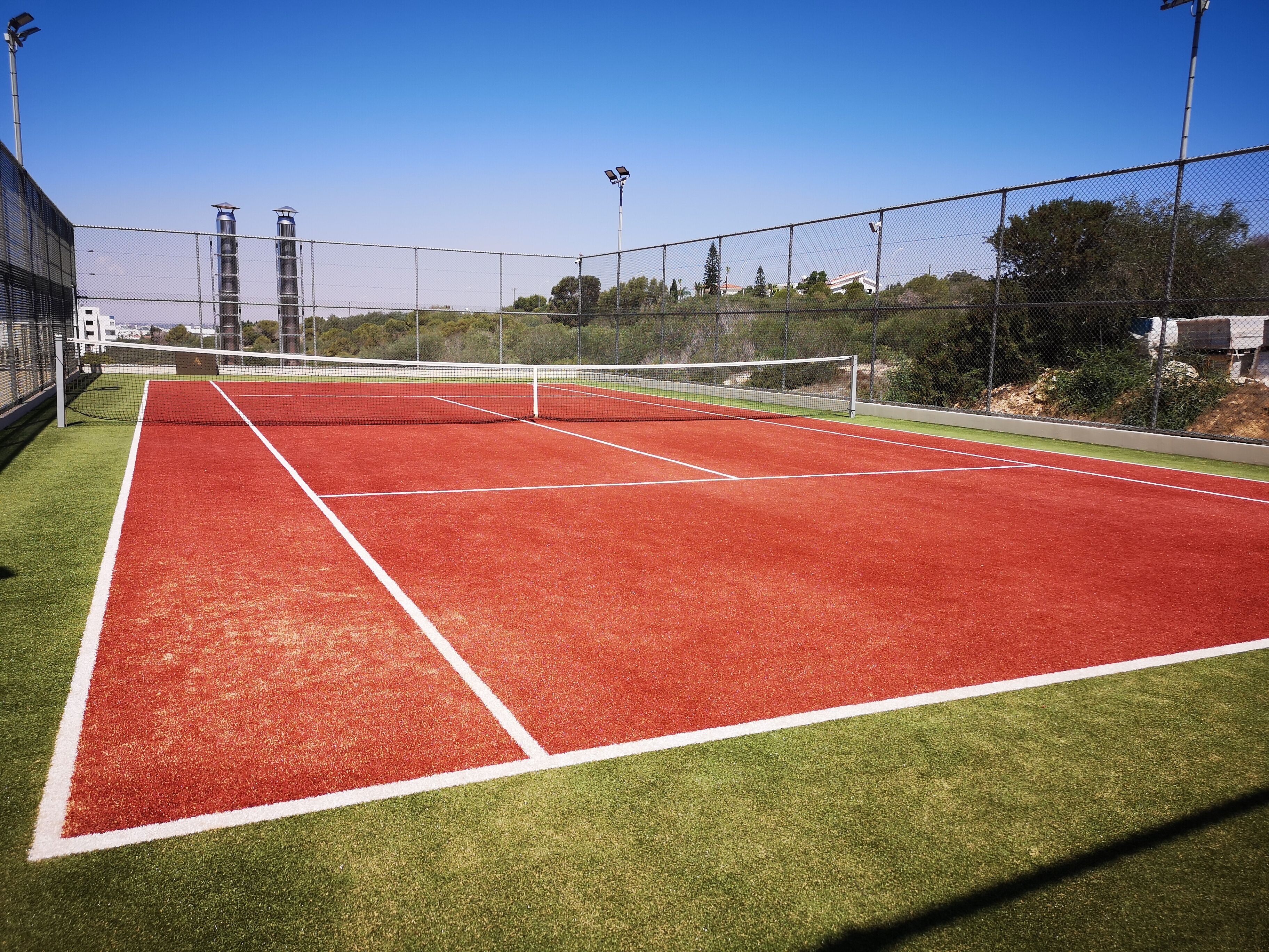 Una cancha de tenis con red, rodeada por una cerca de malla metálica y con iluminación artificial.