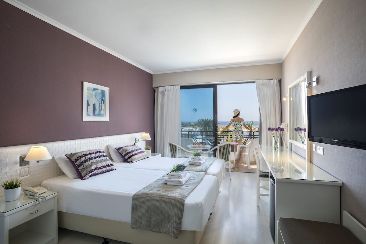 Habitación de hotel con dos camas, un balcón con vistas, televisión y una pequeña mesa con flores.