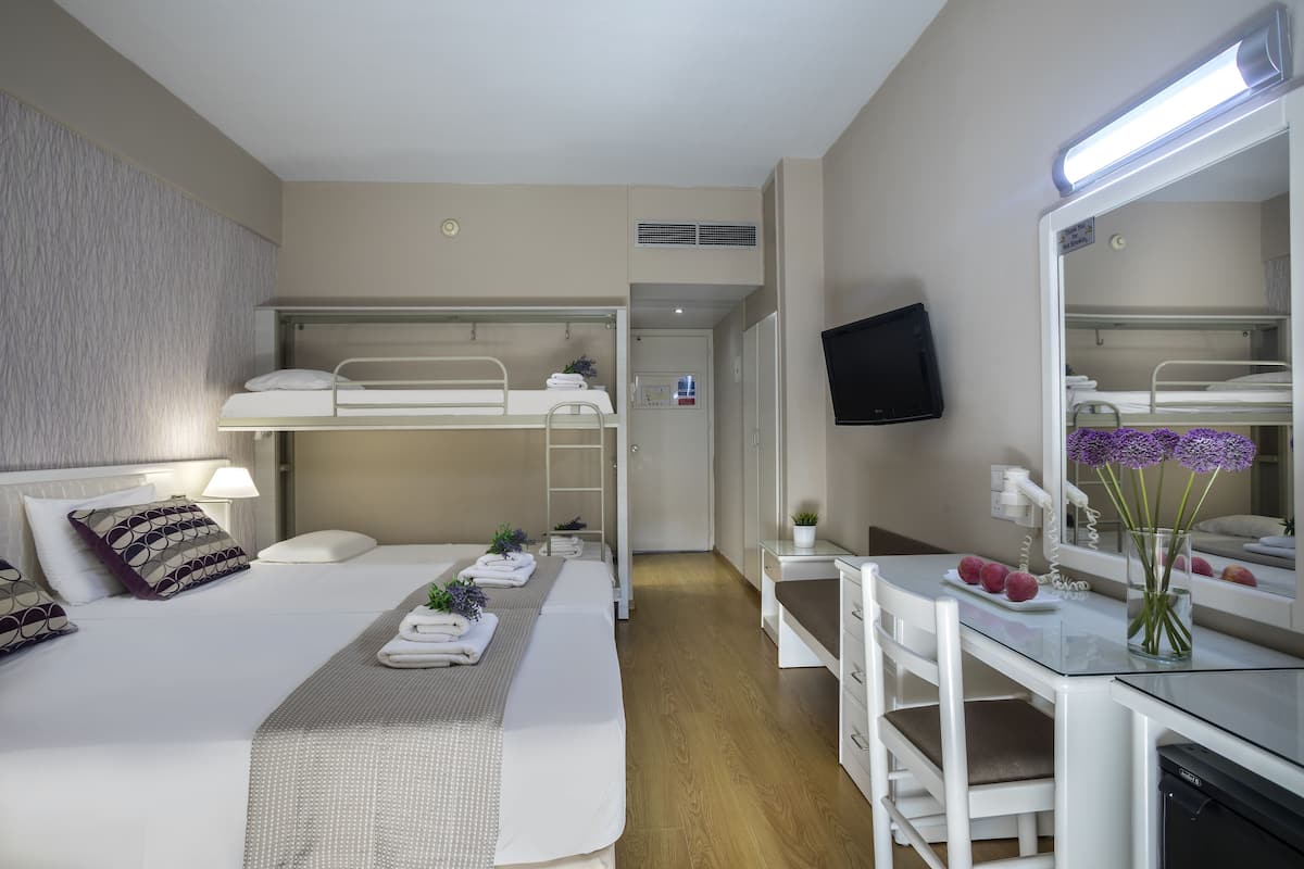 Habitación de hotel con literas, un escritorio con silla, un espejo, un televisor y una pequeña mesa con un jarrón de flores.