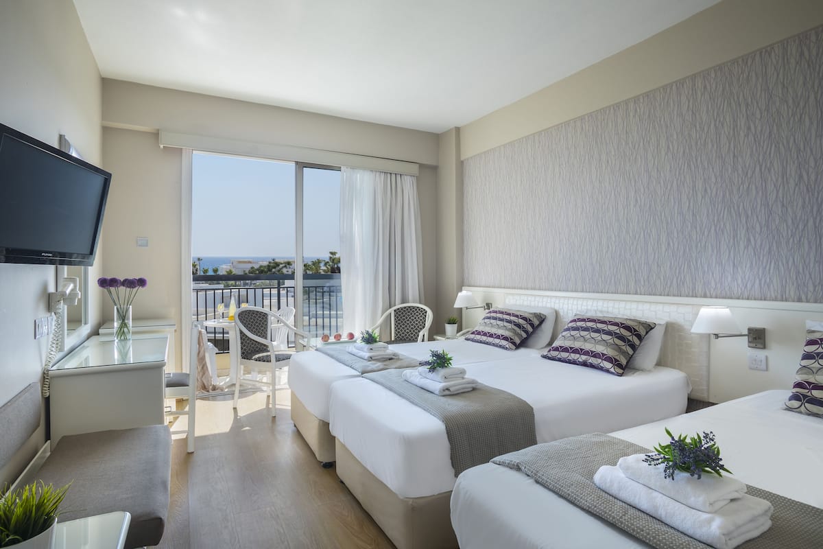 Habitación de hotel con dos camas, un escritorio con televisor, un balcón con vistas y una silla.