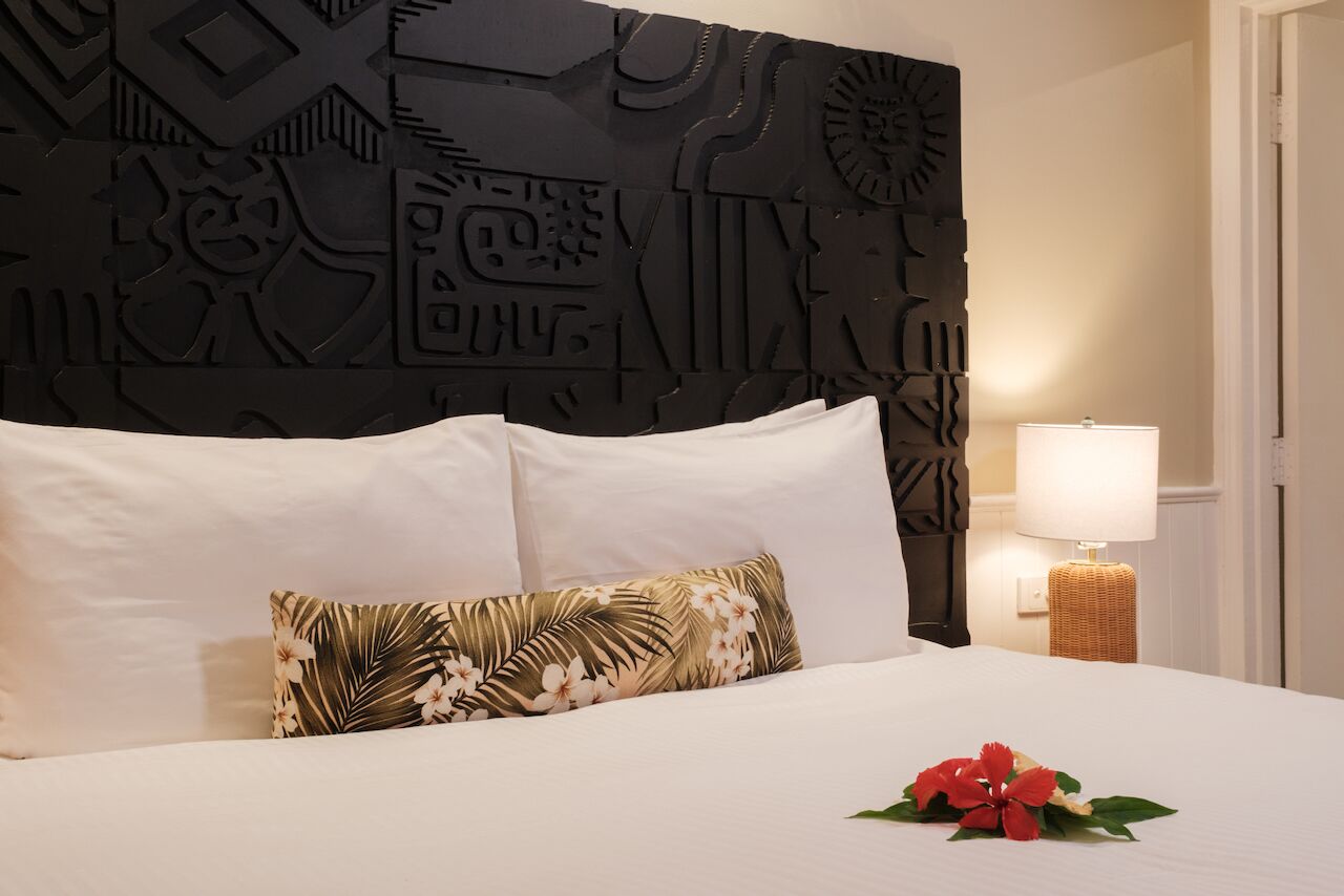 A hotel room with a bed, white pillows, a tropical floral pillow, a decorative headboard, a bedside lamp, and a small table with a lamp.