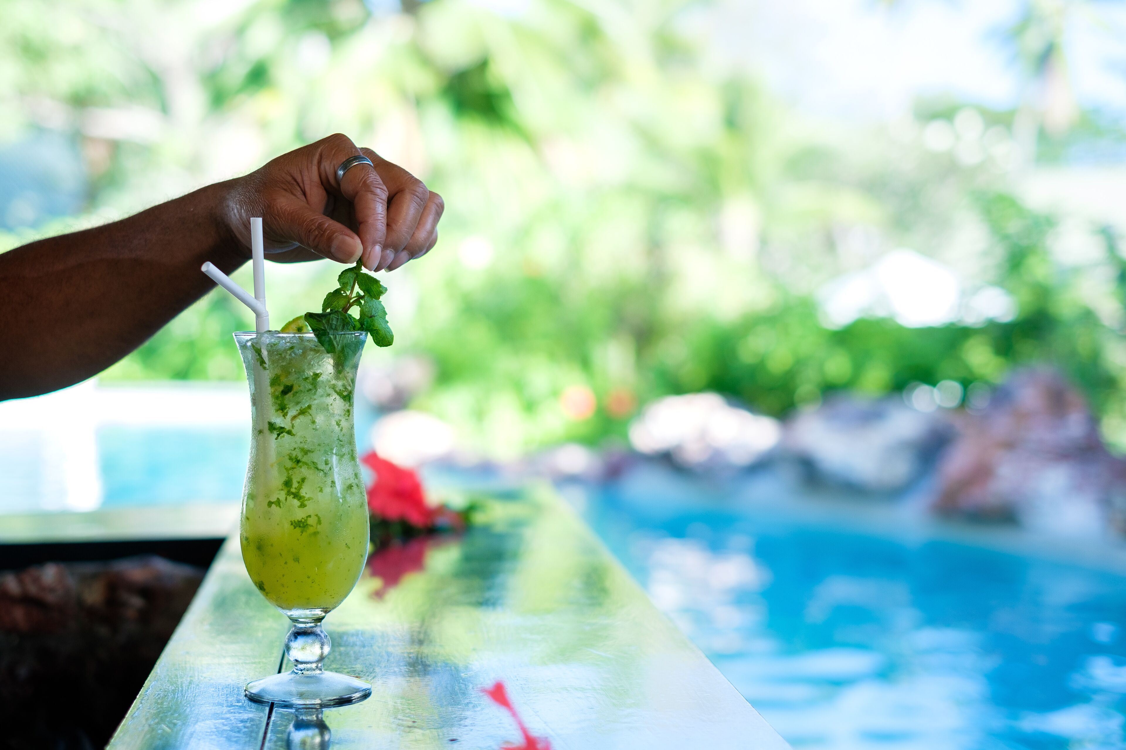 A hand holding a cocktail glass with a straw, placed on a bar counter with a pool in the background.