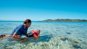 A person in a blue shirt and another in a pink life jacket in clear shallow water with a distant island.