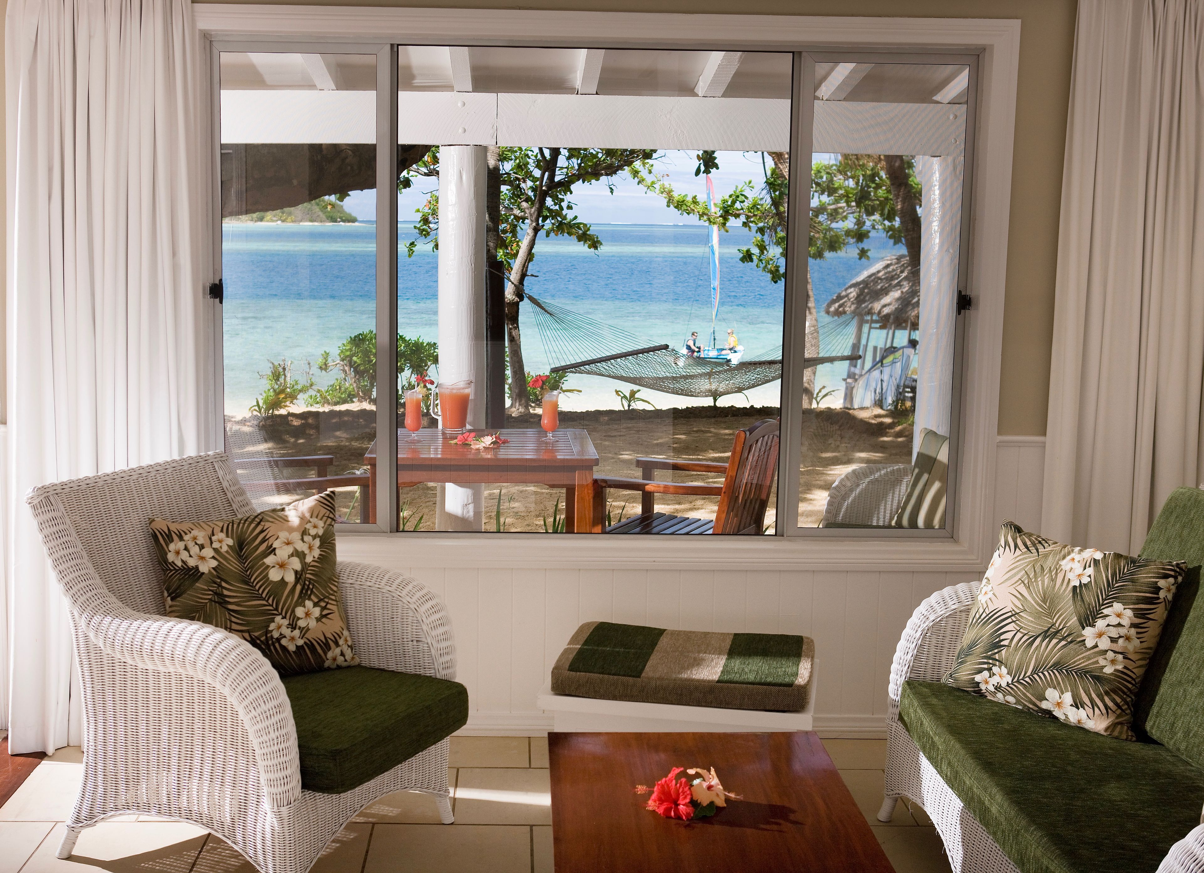 Una habitación con un ventanal grande con vistas a la playa, muebles de mimbre y una mesa de madera.
