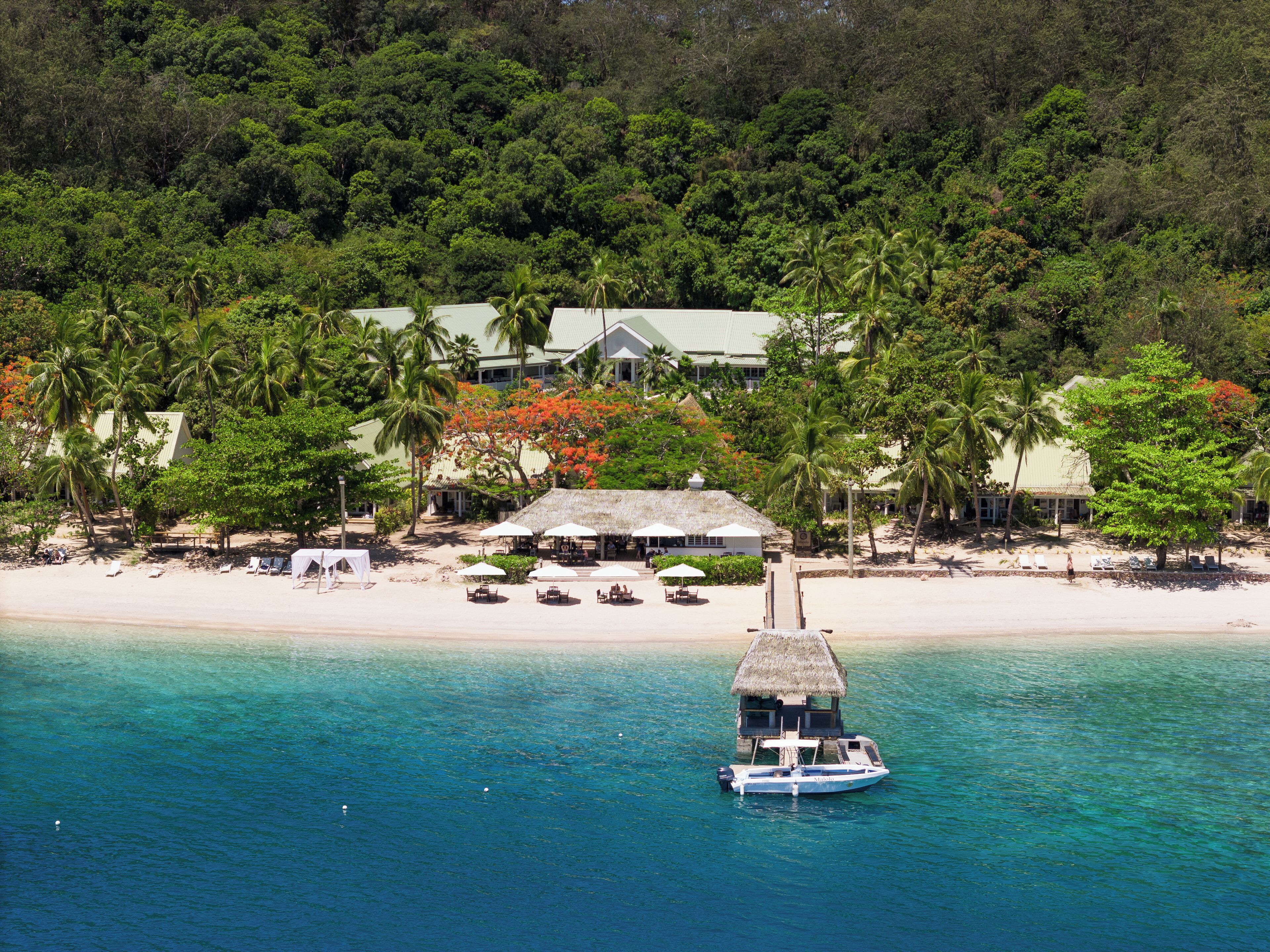A beachfront property with a thatched-roof hut, a boat docked on the shore, and a sandy beach with umbrellas and lounge chairs.