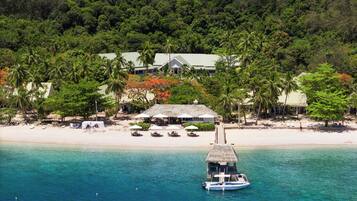 A beachfront property with a thatched-roof hut, a boat docked on the shore, and a sandy beach with umbrellas and lounge chairs.