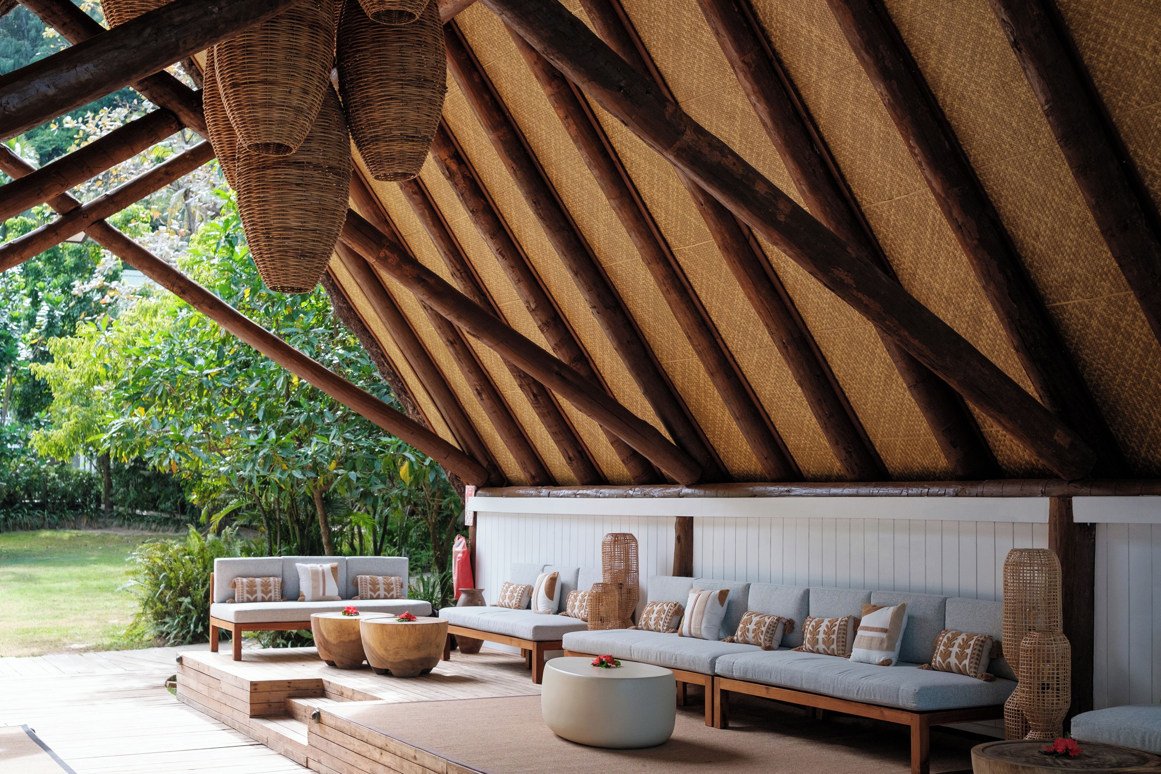 A covered outdoor seating area with a thatched roof, cushioned sofas, and wooden tables.