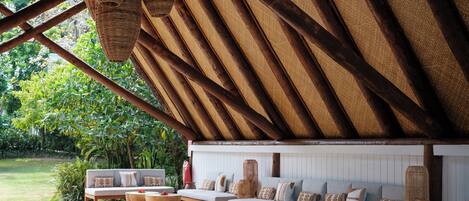 A covered outdoor seating area with a thatched roof, cushioned sofas, and wooden tables.