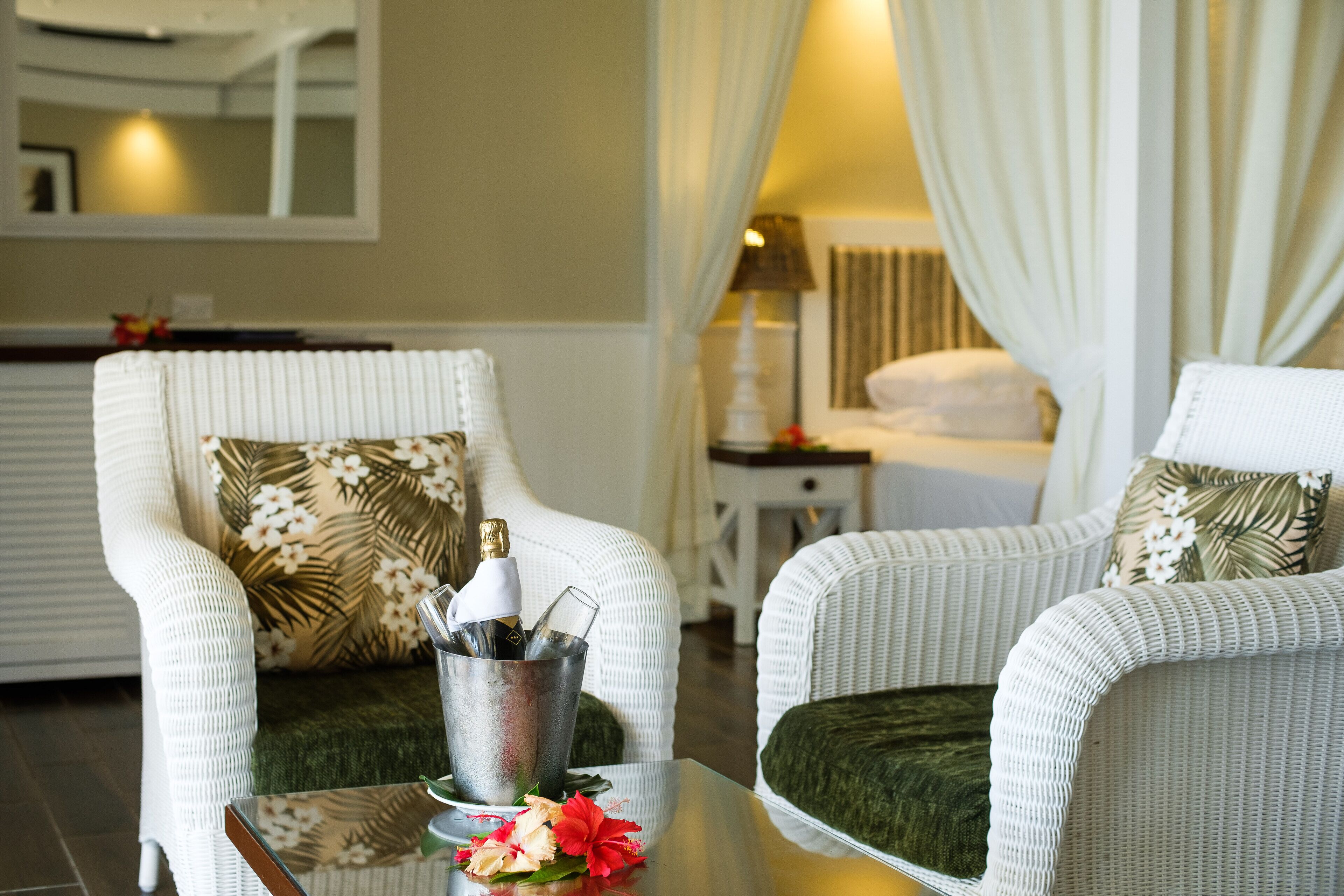 Een hotelkamer met twee witte rieten stoelen, een glazen tafel met een fles en glazen, en een groene kussen.