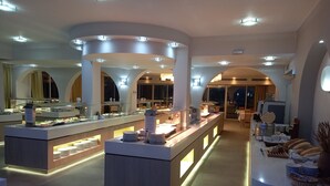 Un moderno bufé de desayuno con varias estaciones de comida, iluminado por luces del techo, y un área de descanso con mesas y sillas.