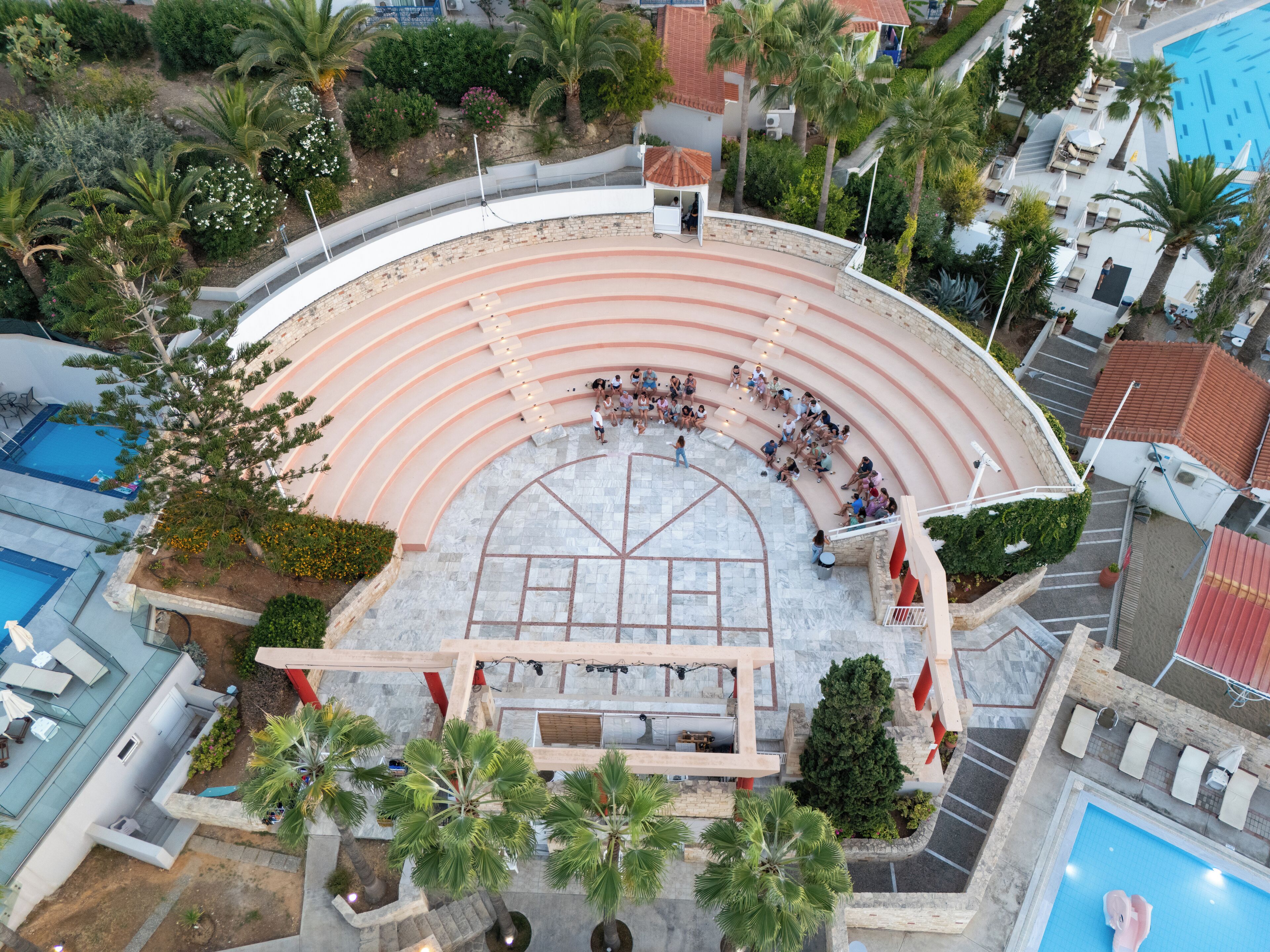 Un anfiteatro al aire libre con asientos escalonados, rodeado de palmeras y una piscina.