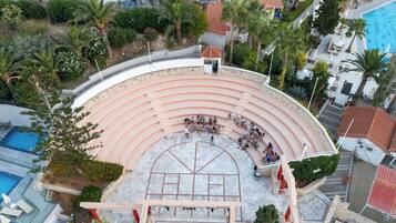 Un anfiteatro al aire libre con asientos escalonados, rodeado de palmeras y una piscina.
