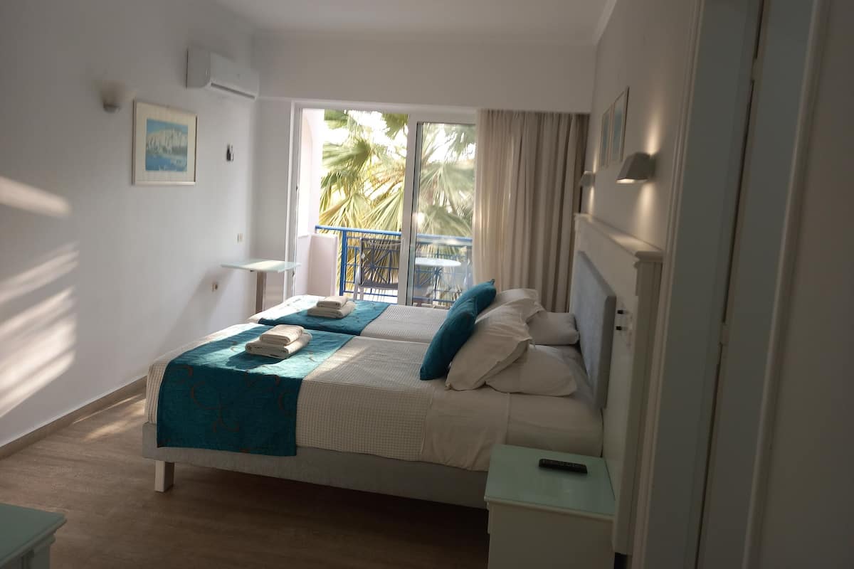 A bedroom with a bed, a nightstand, a small table, a window with curtains, and a balcony with a railing.
