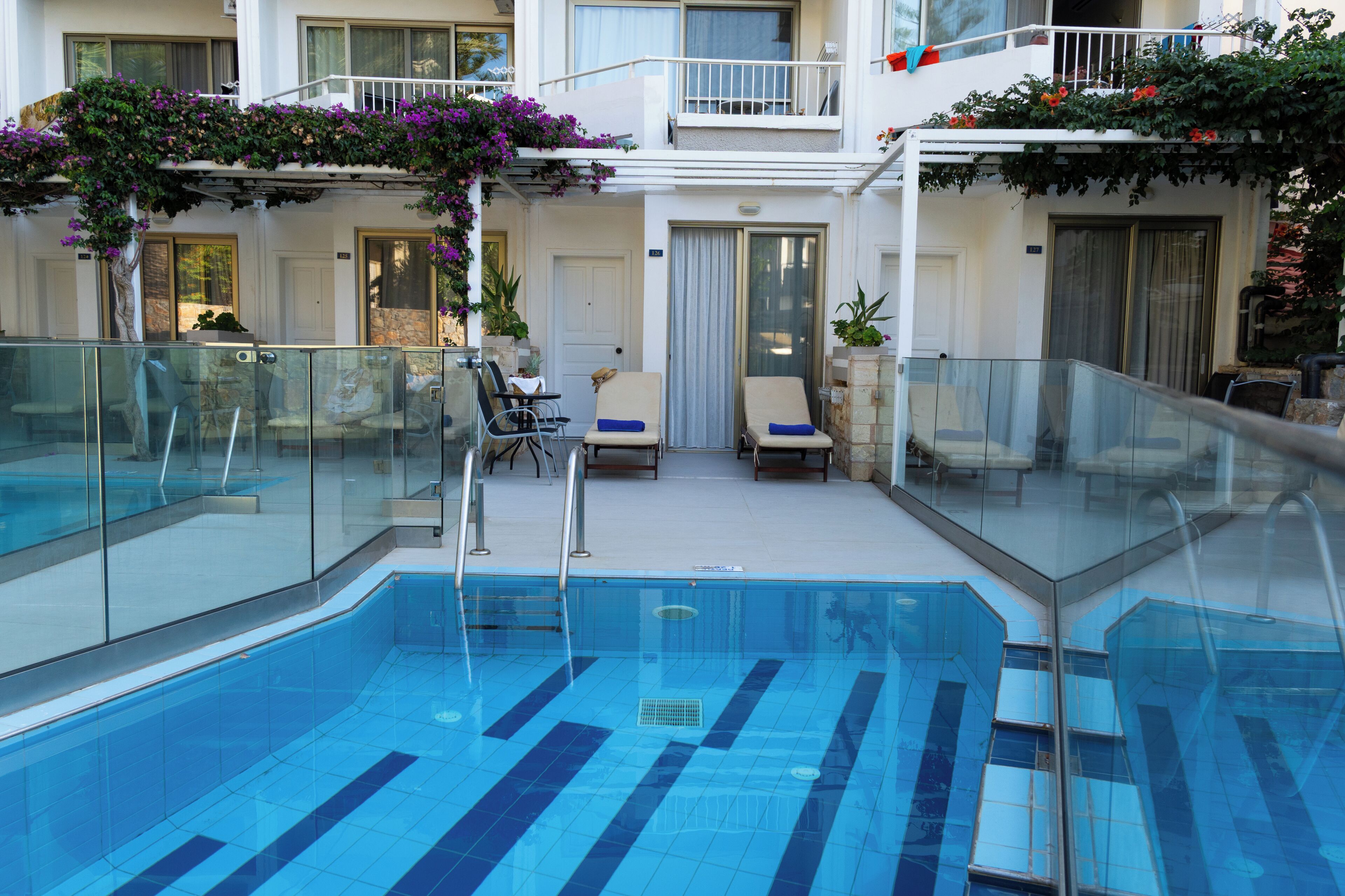 Una piscina con barandillas de vidrio, sillas de descanso y un balcón con plantas.