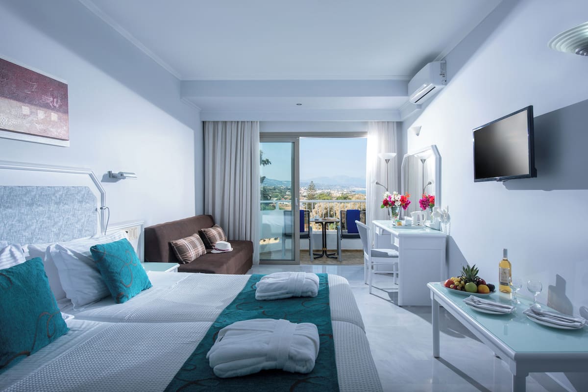 A modern hotel room with a large bed, a desk with a fruit bowl and wine, a balcony with a view, and a flat-screen TV.