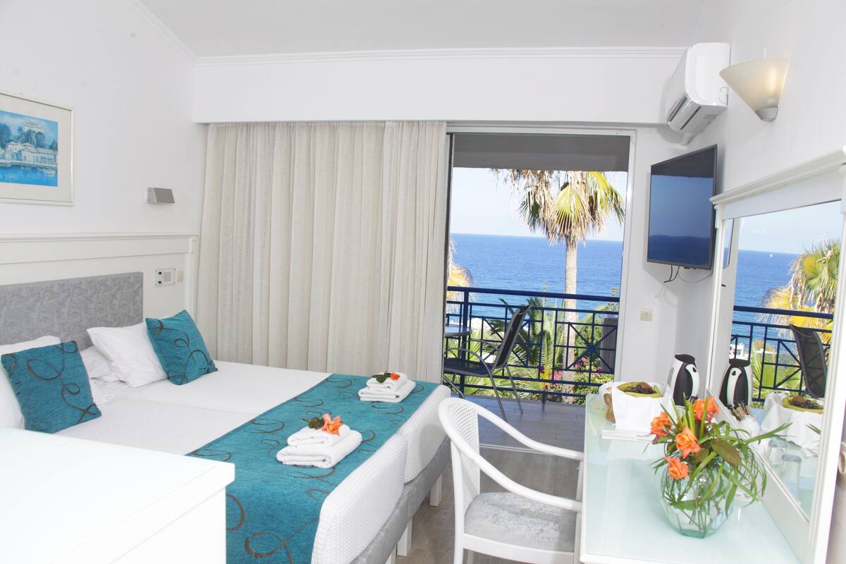 A hotel room with a bed, a desk, a chair, a TV, and a balcony with a view of the sea.