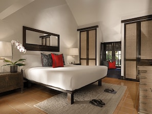 A bedroom with a large bed, a nightstand with a lamp, a potted plant, and a view of the outside through a door.