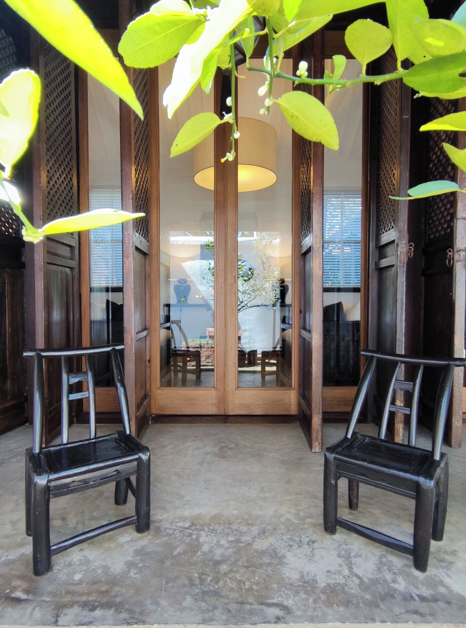 Una habitación con dos sillas de madera, una mesa y vistas a un área exterior con plantas y una estructura blanca.
