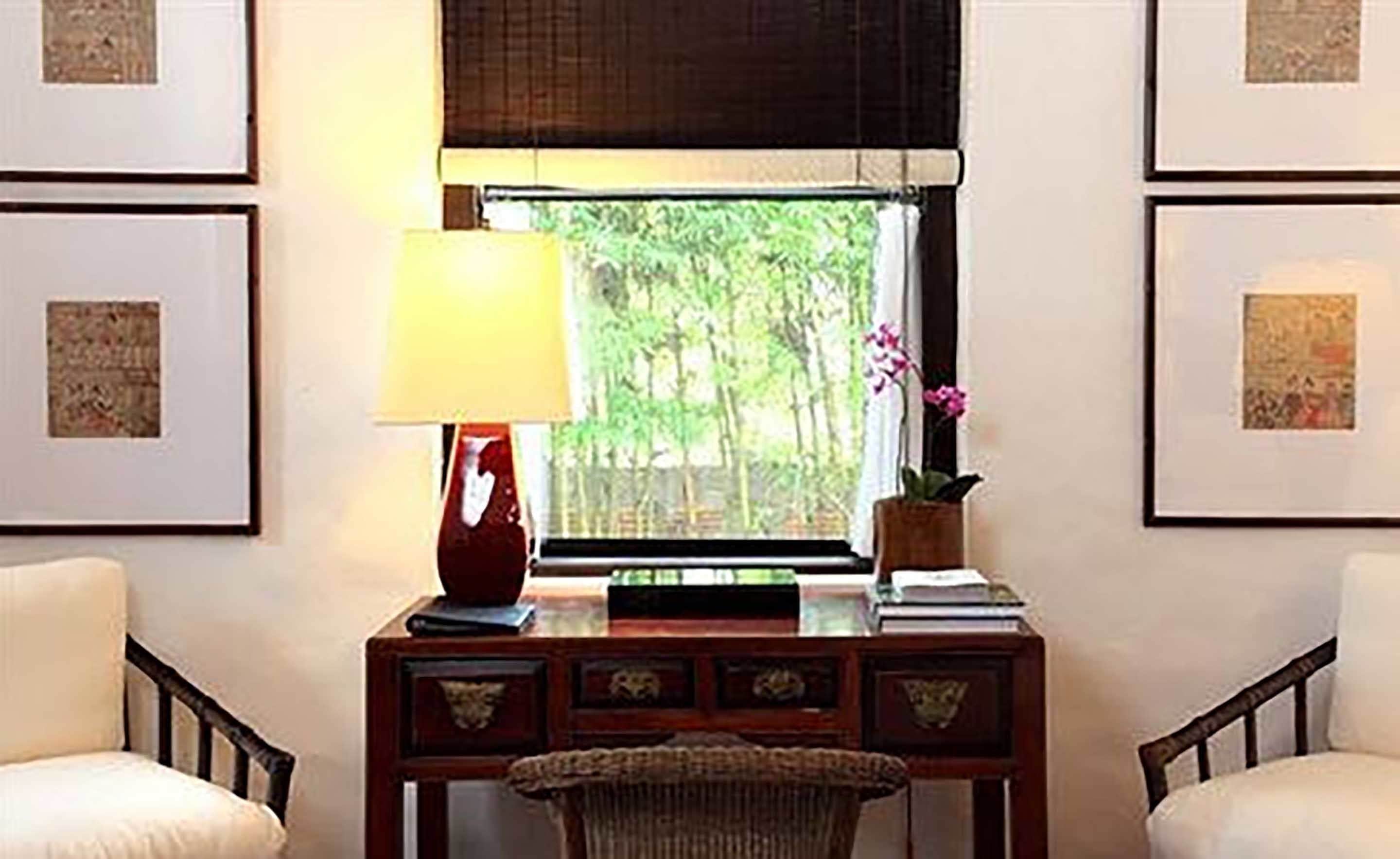 A room with a desk, a lamp, a window with a view of greenery, and framed pictures on the walls.
