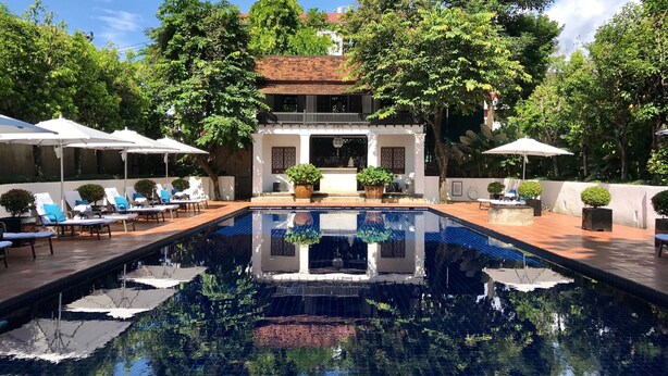A pool area with lounge chairs and umbrellas, surrounded by trees and a building with a tiled roof. - Rachamankha Hotel (Chiang Mai)